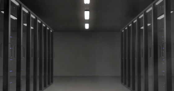 Serwerownia w ciemnej kolorystyce - po lewej i prawej ustawione szafy serwerów, na środku pusta przestrzeń, na górze sufit ze świecącymi lampami